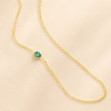 A yellow gold 14K Gold Asymmetrical Birthstone Necklace - Emerald (May) on a beige background.