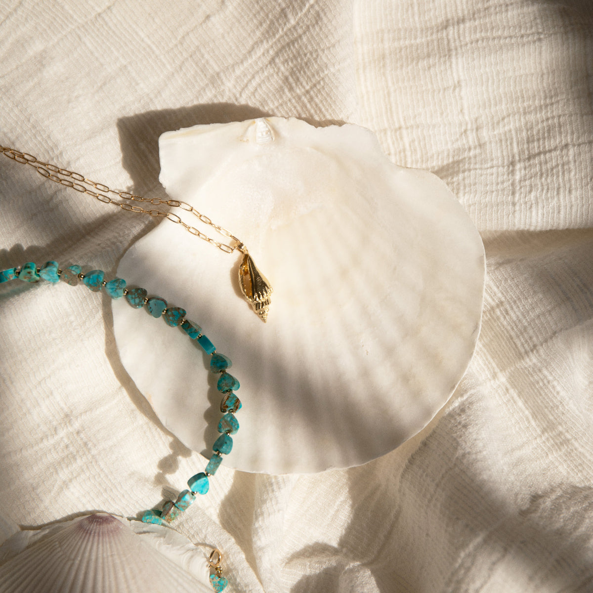 An Endless Summer Necklace - Sea + Sky and Element Short Link Chain with a Golden Spiral Shell on a white shell and white textured fabric. 