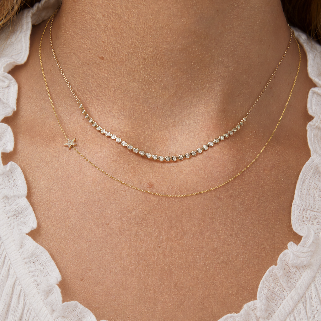 Close-up of a person's neck wearing The Everyday Diamond Necklace layered with a 14k Gold Asymmetrical Charm Necklace - Pavé Star in yellow gold, styled with a cream top.
