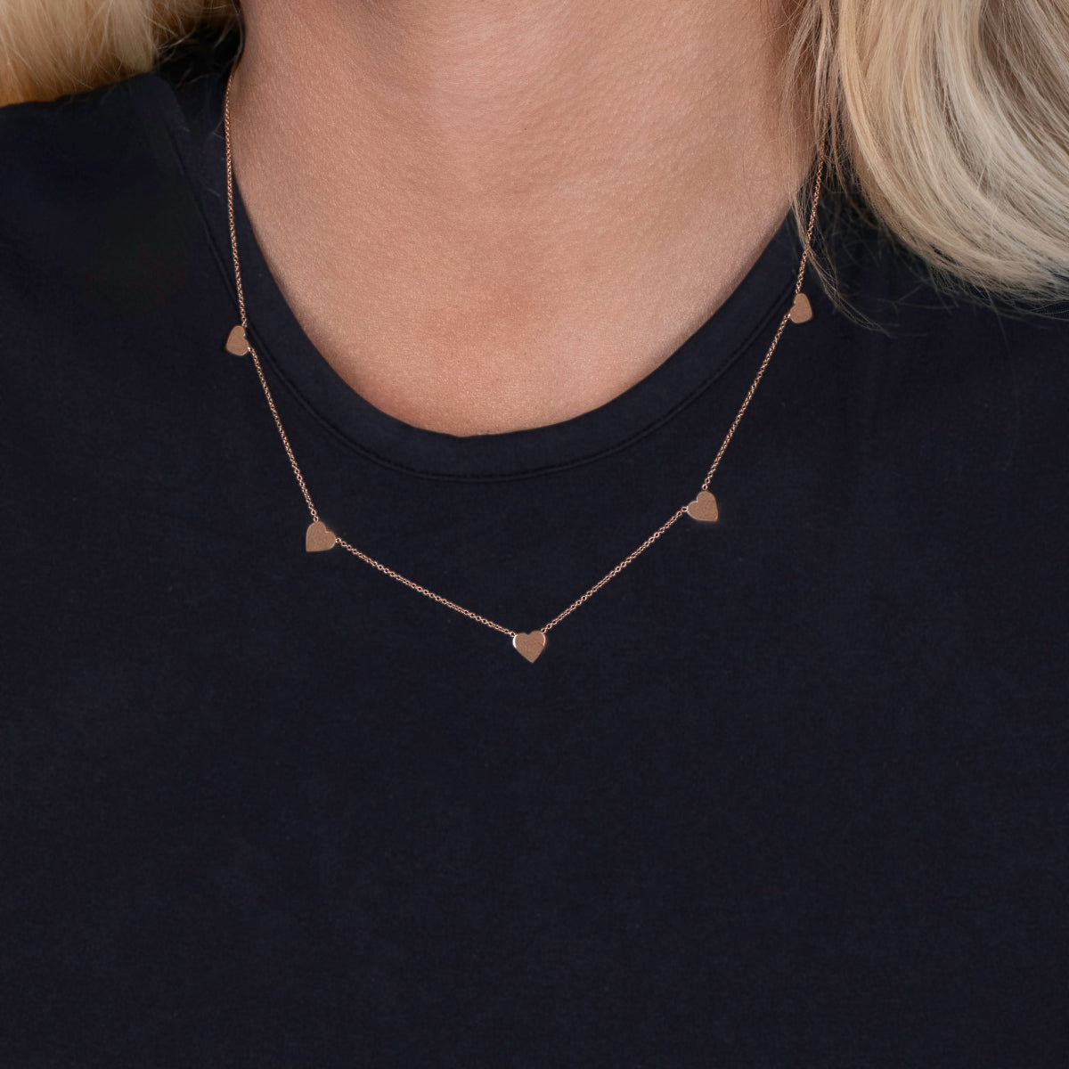 A close-up of a woman's décolletage wearing a 5 of Hearts Necklace with a black top and her blonde hair partially visible. 