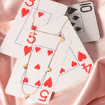 A rose gold 5 of Hearts Necklace on top of playing cards and a pink silk fabric background. 