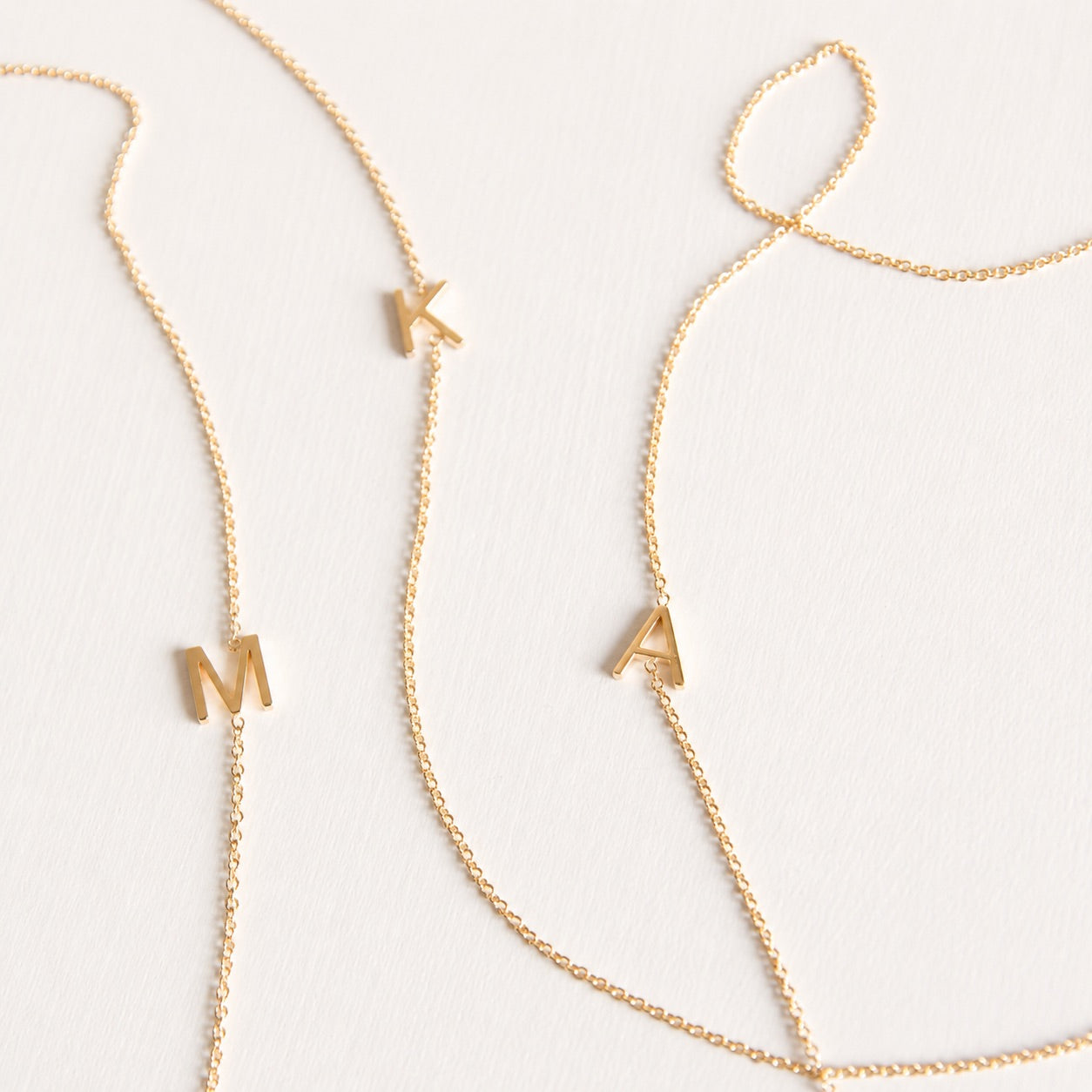 Three gold necklaces with single letter charms, being M, K and A on a plain background. 