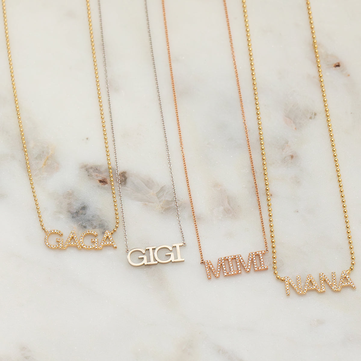 A Pavé GAGA Necklace in yellow gold, a GIGI Necklace in white gold, a Pavé MIMI Necklace in rose gold, and a Pavé NANA Necklace in yellow gold, displayed on a light marble surface.