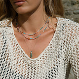 Close-up of a blonde woman’s décolletage wearing two Endless Summer Necklaces - Capri and The Inflow Retreat Necklace,  styled in a cream crochet top against a stone-blurred background.