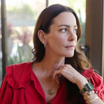 A brunette woman wearing a yellow gold Be Gentle in Grief Necklace, along with several earrings, bracelets and a watch, dressed in a red shirt inside a house with the backyard visible in the background.
