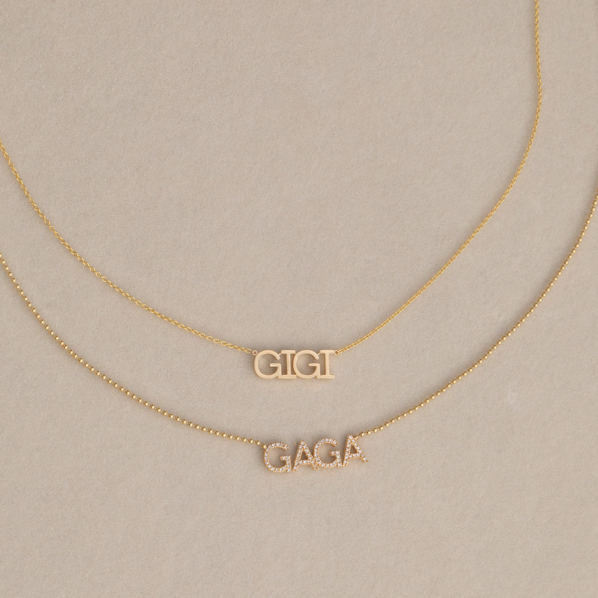 A GIGI Necklace in yellow gold with a cable chain and a Pavé Necklace in yellow gold with a bead chain, against a light beige background. 