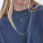 A close-up of a blonde woman's décolletage wearing a New York pendant on a chain necklace and an Overboard Layering Necklace, styled with a blue jumper against a white background. 