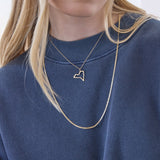 A close-up of a blonde woman's décolletage wearing a New York pendant on a chain necklace and an Overboard Layering Necklace, styled with a blue jumper against a white background. 