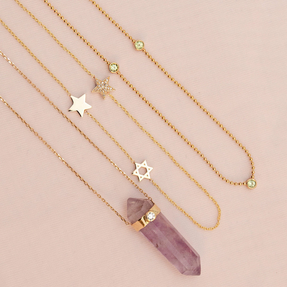 The Healing Retreat Necklace, a Custom Gold Necklace - 3 Letters with star charms and a Triple Birthstone Layering Necklace - Yellow Gold (Peridot) laid on a light pink background. 