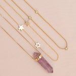 The Healing Retreat Necklace, a Custom Gold Necklace - 3 Letters with star charms and a Triple Birthstone Layering Necklace - Yellow Gold (Peridot) laid on a light pink background. 