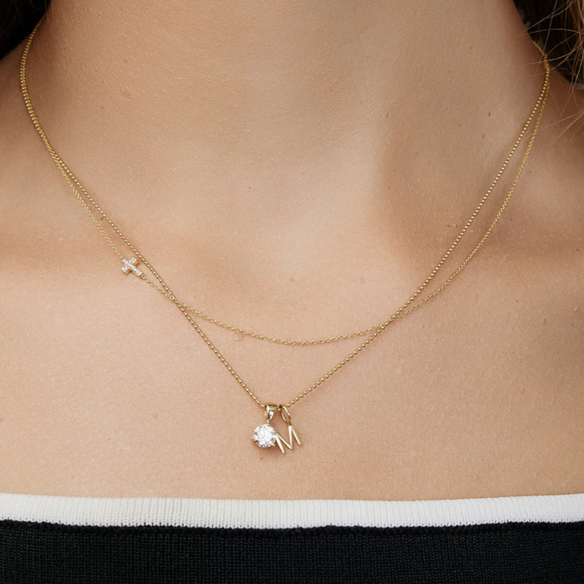 Close-up of a neck wearing a Classic Bead Chain with a Gold Letter Charm and Duchess Diamond Pendant, and a 14k Gold Asymmetrical Charm Necklace - Pave Cross, both in yellow gold. 