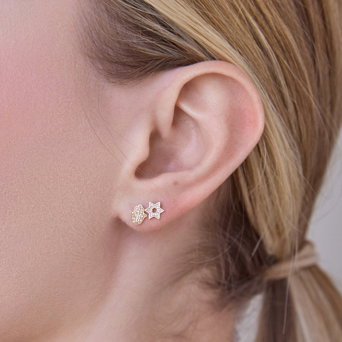 Close-up of a woman's ear wearing Pavé Charm Stud - Hamsa in yellow gold and Pavé Charm Stud - Star of David in white gold, with her blonde hair visible. 