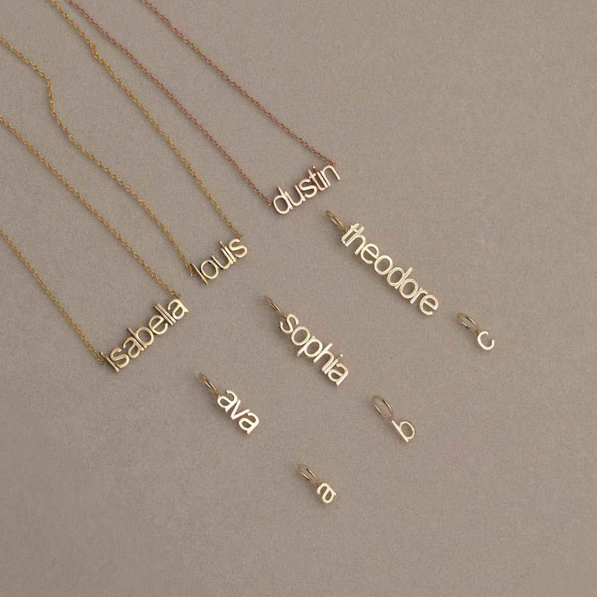 Three Petite Name Plate Necklaces with the names Isabella, Louis and Dustin, three Petite Nameplate Pendants in the names Ava, Sophia and Theodore, and three Petite Letter Charms in lowercase a, b and c, all in yellow gold, against a beige background. 