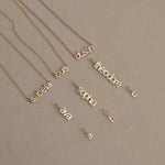 Three Petite Name Plate Necklaces with the names Isabella, Louis and Dustin, three Petite Nameplate Pendants in the names Ava, Sophia and Theodore, and three Petite Letter Charms in lowercase a, b and c, all in yellow gold, against a beige background. 