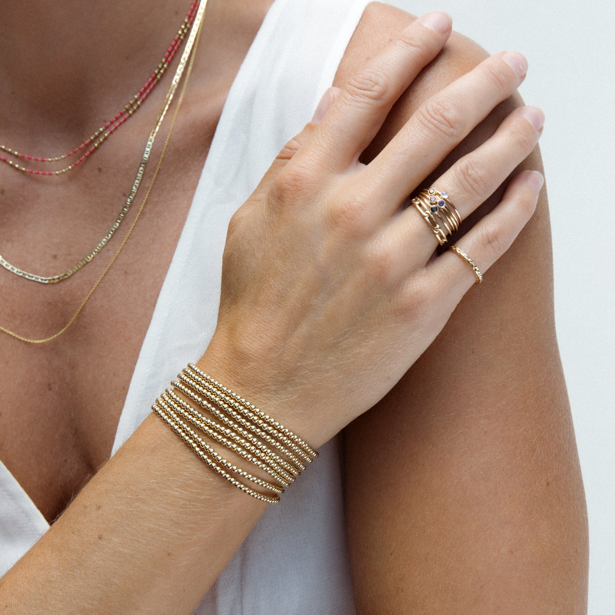 A close-up of a woman's hand resting on her shoulder, wearing multiple stacked Little Bubble Bracelets, a Link Ring, multiple stacked Birthstone Rings, a Braided Ring and Confetti Necklaces in pink and green, with a white top. 
