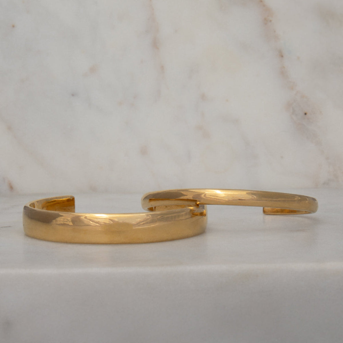 An All In Cuff and a Plus One Cuff in vermeil gold on a marble surface and background.
