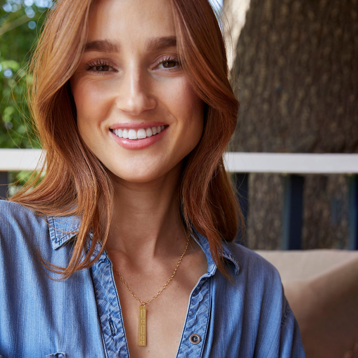 A woman wearing a Virtues Pendant - Rectangle in yellow gold with a blue denim shirt against an outdoors blurred background. 
