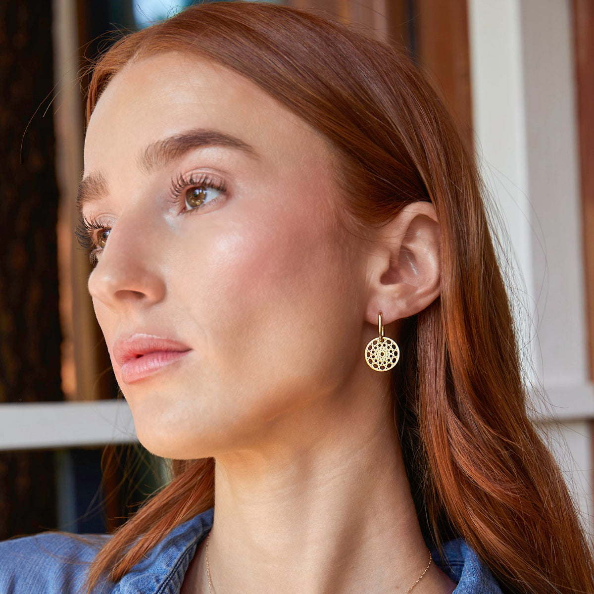 Close-up of a woman wearing a gold Skylight Hoop with orange hair and a denim blue top against a blurred background. 