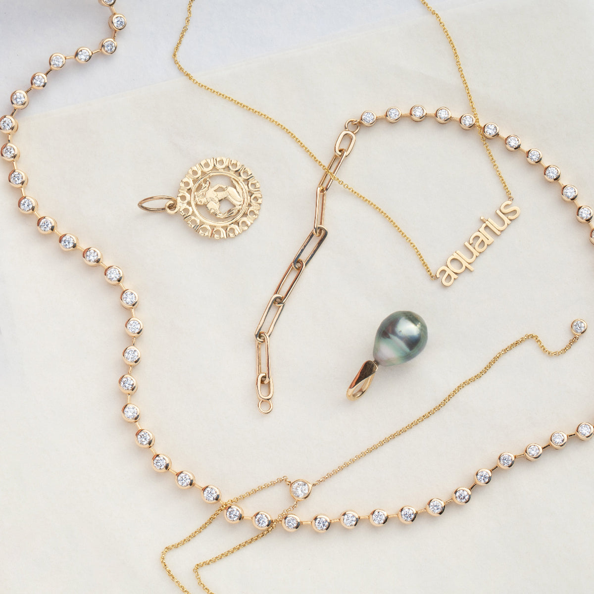 The Ultimate Diamond Necklace, a Petite Zodiac Necklace 'aquarius', a Diamond Drip Necklace, a Zodiac Pendant and a Baroque Pearl Pendant laid on a cream background. 