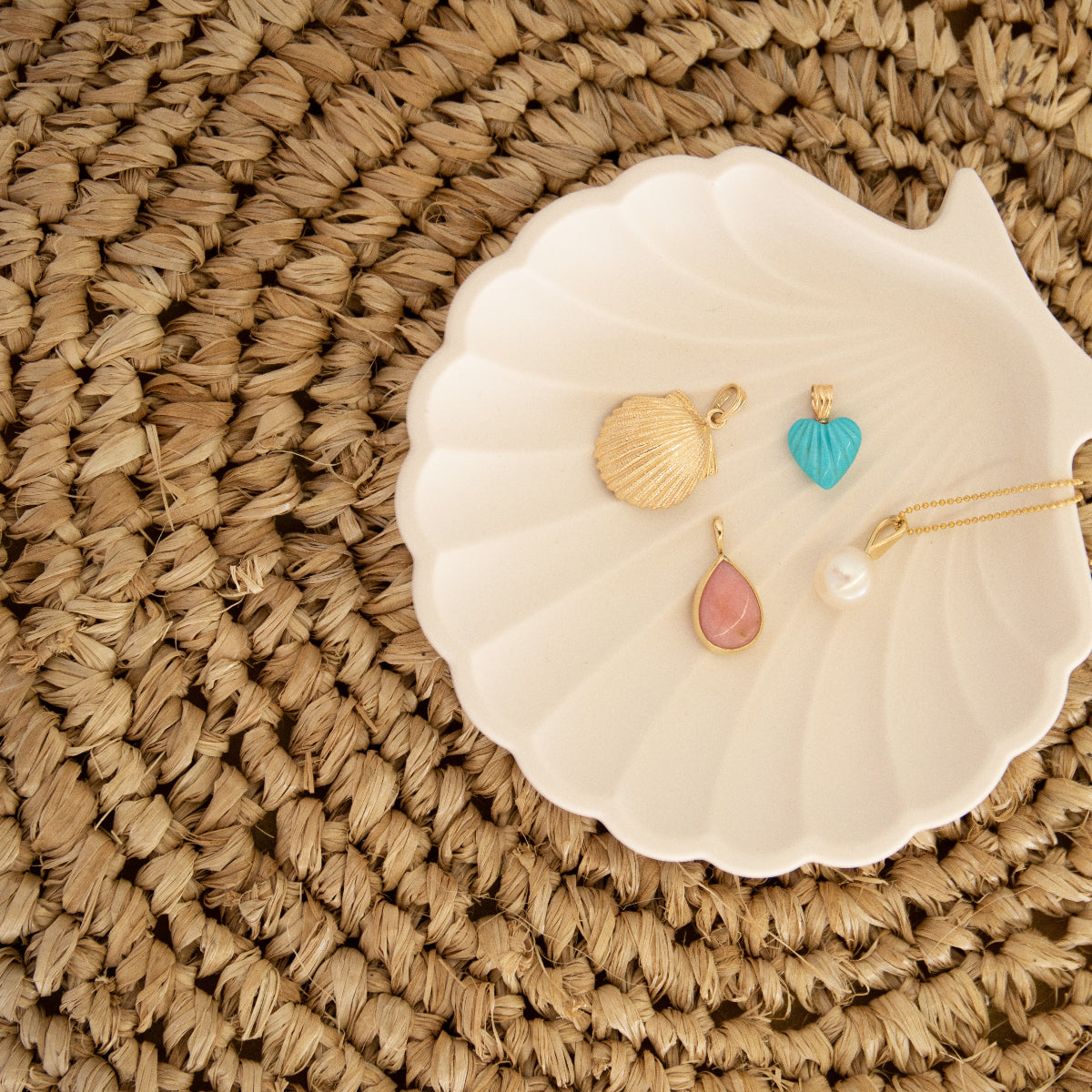 A Golden Scallop Shell, Etched Turquoise Heart Charm, Pink Opal Tear Drop Charm  and a Perla Necklace in a white shell holder against a textured woven surface. 