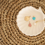 A Golden Scallop Shell, Etched Turquoise Heart Charm, Pink Opal Tear Drop Charm  and a Perla Necklace in a white shell holder against a textured woven surface. 