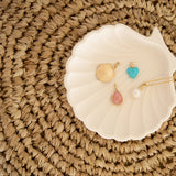 A Golden Scallop Shell, Etched Turquoise Heart Charm, Pink Opal Tear Drop Charm  and a Perla Necklace in a white shell holder against a textured woven surface. 