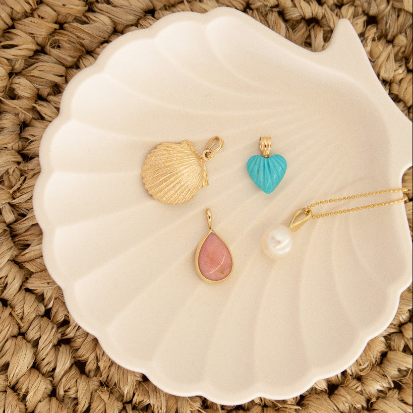 A Golden Scallop Shell, Etched Turquoise Heart Charm, Pink Opal Tear Drop Charm  and a Perla Necklace in a white shell holder against a textured woven surface. 