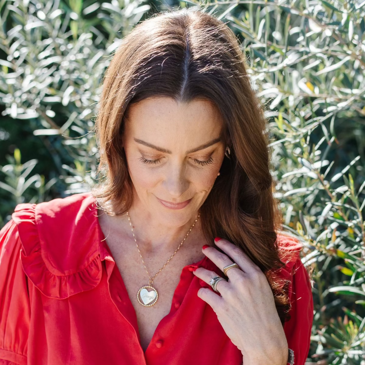 A brunette woman wearing a yellow gold Be Gentle in Grief Necklace along with several rings, including a Geo Stacking Ring Square, dressed in a red shirt against a green foliage background. 