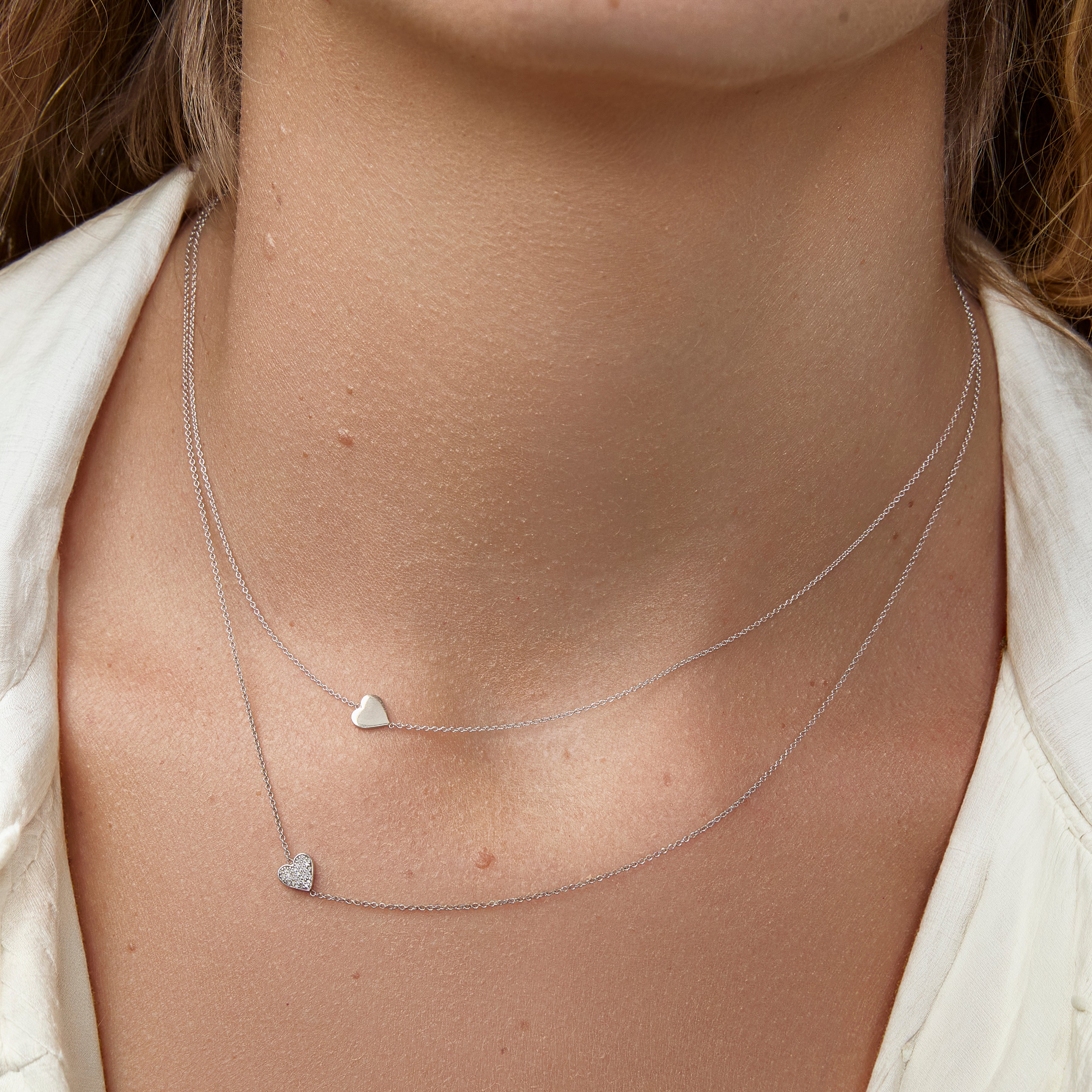 A close-up of a woman's neck wearing two white gold 14k Gold Asymmetrical Charm Necklaces in - Heart and Pavé Heart, styled in a white jacket.