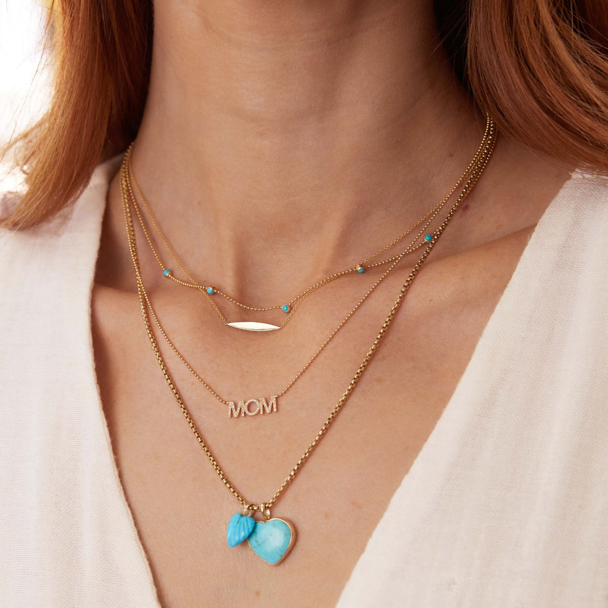 A close-up of a woman's neck wearing a Quintet Birthstone Layering Necklace - Yellow Gold in Turquoise, a Horizon Necklace, a Pavé MUM Necklace, a Box Chain with a Turquoise Heart Charm and an Etched Turquoise Heart Charm, with orange hair styled in a white top.