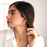 A woman wearing a Sleek & Chic Hoop - Large with two necklaces  in yellow gold and a white shirt, with brown hair against a light background. 