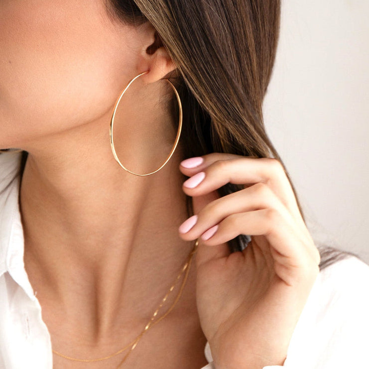 A woman wearing a Sleek & Chic Hoop - Large with two necklaces  in yellow gold and a white shirt, with brown hair against a light background. 