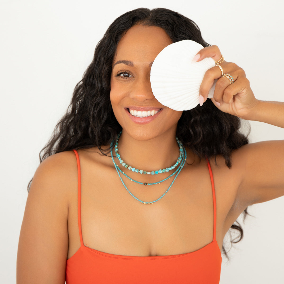 A woman wearing Endless Summer Necklaces in Turqs + Caicos, Santorini, and Sea + Sky, along with several Bouquet Rings and a red strap top, holding a white shell over her face against a white background.