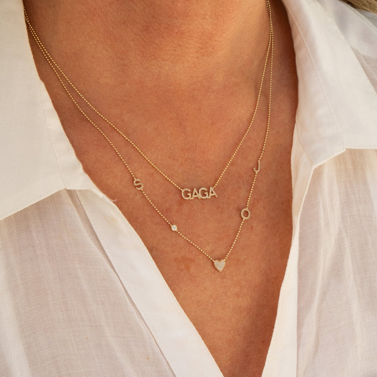 Close-up of a neck wearing a Pavé GAGA Necklace and a Custom Gold Necklace - 5 Letters, both in yellow gold, with a white shirt.