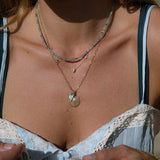 Close-up of a woman's neck wearing an Endless Summer Necklace - Blue Lagoon, a Custom Gold Necklace and an Element Short Link Chain with an Abalone Leaf Charm and a Golden Scallop Shell, styled in a light blue strap top. 