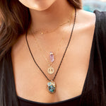 A close-up of a woman's décolletage wearing a yellow gold Custom Gold Necklace - 4 Letters, The Healing Retreat Necklace, a Zodiac Necklace - Gemini and  a Black Rhodium Antique Bead Chain with a Large Abalone Cabochon Charm, Abalone Leaf Charm and a Small Abalone Cabochon Charm, with brown hair and a black top. 