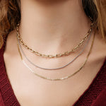 Close-up of a woman's décolletage wearing a Bold Link Chain in yellow gold, a white gold Box Chain and a yellow gold Overboard Layering Chain, with a dark red top and her blonde hair visible. 