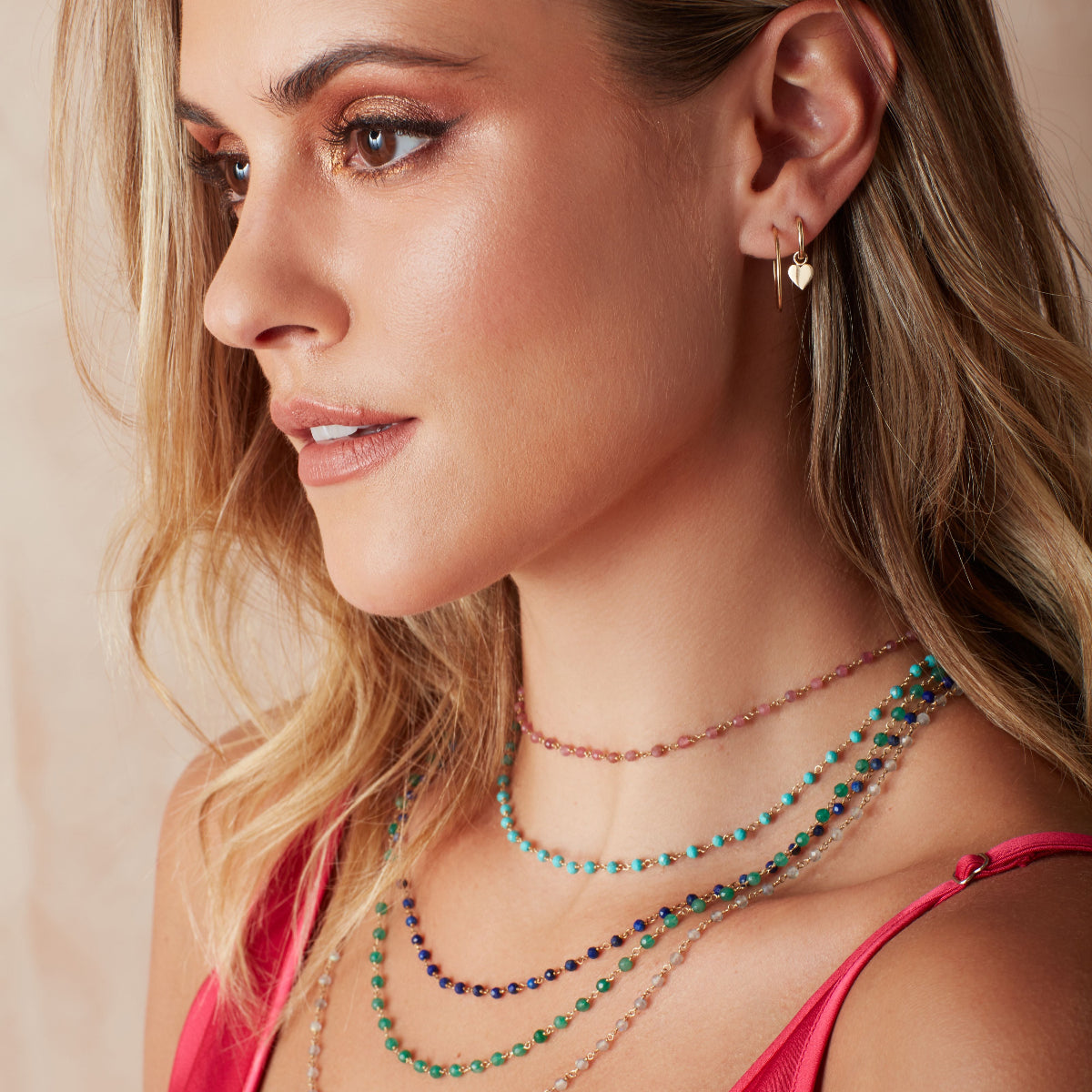 A blonde woman wearing The Peony Bouquet Necklace, The Bluebell Bouquet Necklace, The Thistle Bouquet Necklace, The Closer Bouquet Necklace, The Sage Bouquet Necklace, a Charmed I'm Sure Hoop / Heart and a Twisted Eclipse Stud, with a dark pink top against a neutral background. 