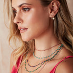 A blonde woman wearing The Peony Bouquet Necklace, The Bluebell Bouquet Necklace, The Thistle Bouquet Necklace, The Closer Bouquet Necklace, The Sage Bouquet Necklace, a Charmed I'm Sure Hoop / Heart and a Twisted Eclipse Stud, with a dark pink top against a neutral background. 