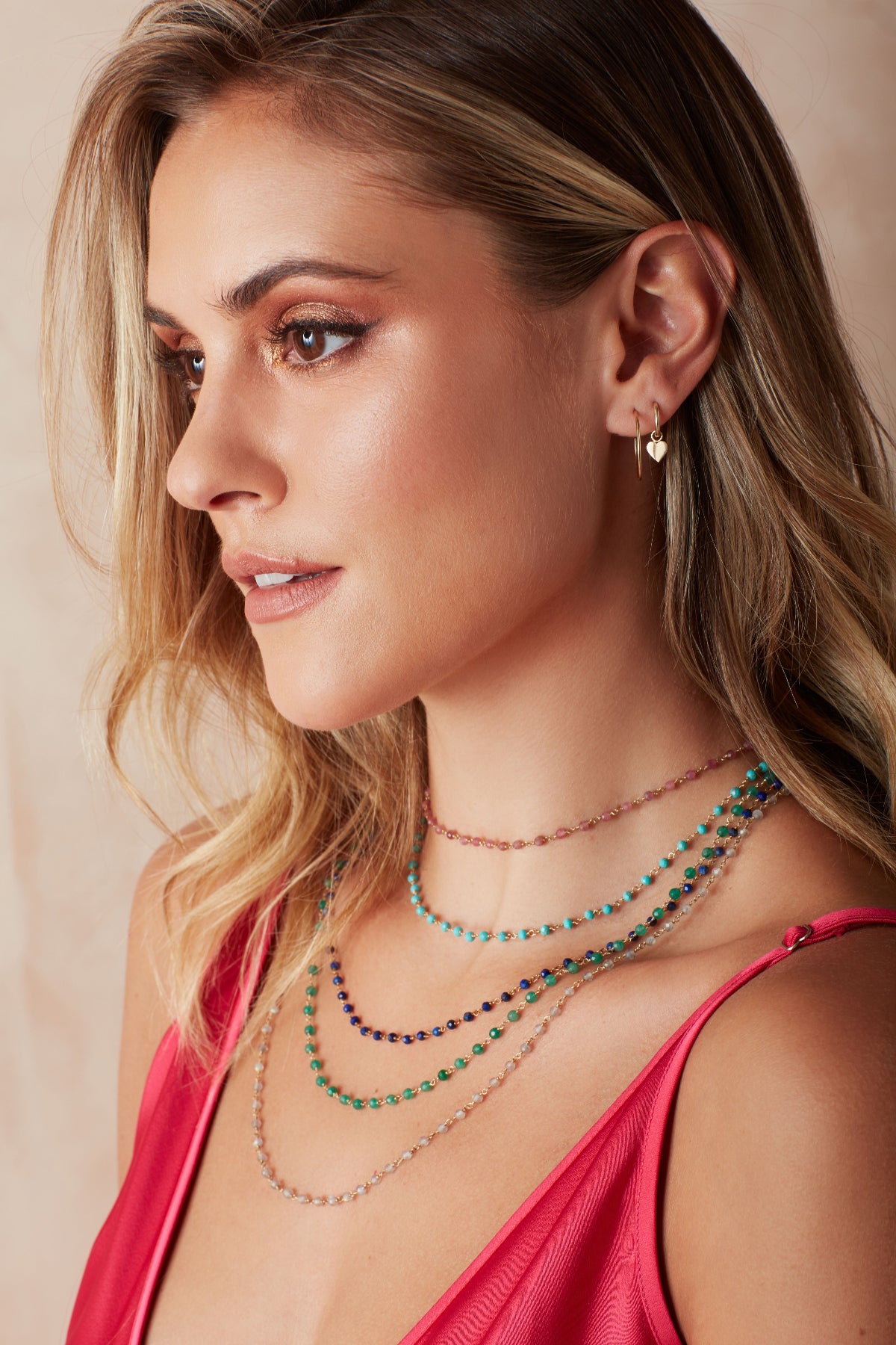 A blonde woman wearing The Peony Bouquet Necklace, The Bluebell Bouquet Necklace, The Thistle Bouquet Necklace, The Closer Bouquet Necklace, The Sage Bouquet Necklace, a Charmed I'm Sure Hoop / Heart and a Twisted Eclipse Stud, with a dark pink top against a neutral background. 