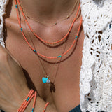 Close-up of a neck wearing a yellow gold Custom Gold Necklace, Endless Summer Necklaces and Bracelets - Calypso and Mykonos, a yellow gold Classic Bead Chain with an Etched Turquoise Heart Charm, a Diamond Pendant and a Petite Nameplate Pendant, styled in a navy blue bikini top and a white crochet top.