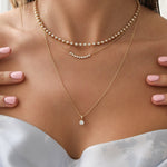 Close-up of a woman's décolletage wearing The Ultimate Diamond Necklace, A Diamond Arc Layering Necklace and a Classic Bead Chain with a Princess Diamond Pendant, all in yellow gold, with a light grey and white floral top.
