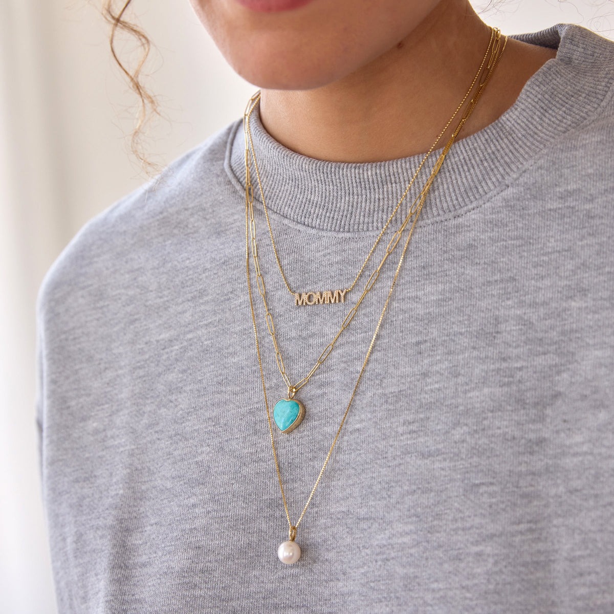 A close-up of a person wearing a pave MOMMY Necklace, an Element Long Link Chain with a Turquoise Heart Charm and a Perla Necklace, styled in a light grey jumper. 