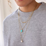 A close-up of a person wearing a pave MOMMY Necklace, an Element Long Link Chain with a Turquoise Heart Charm and a Perla Necklace, styled in a light grey jumper. 