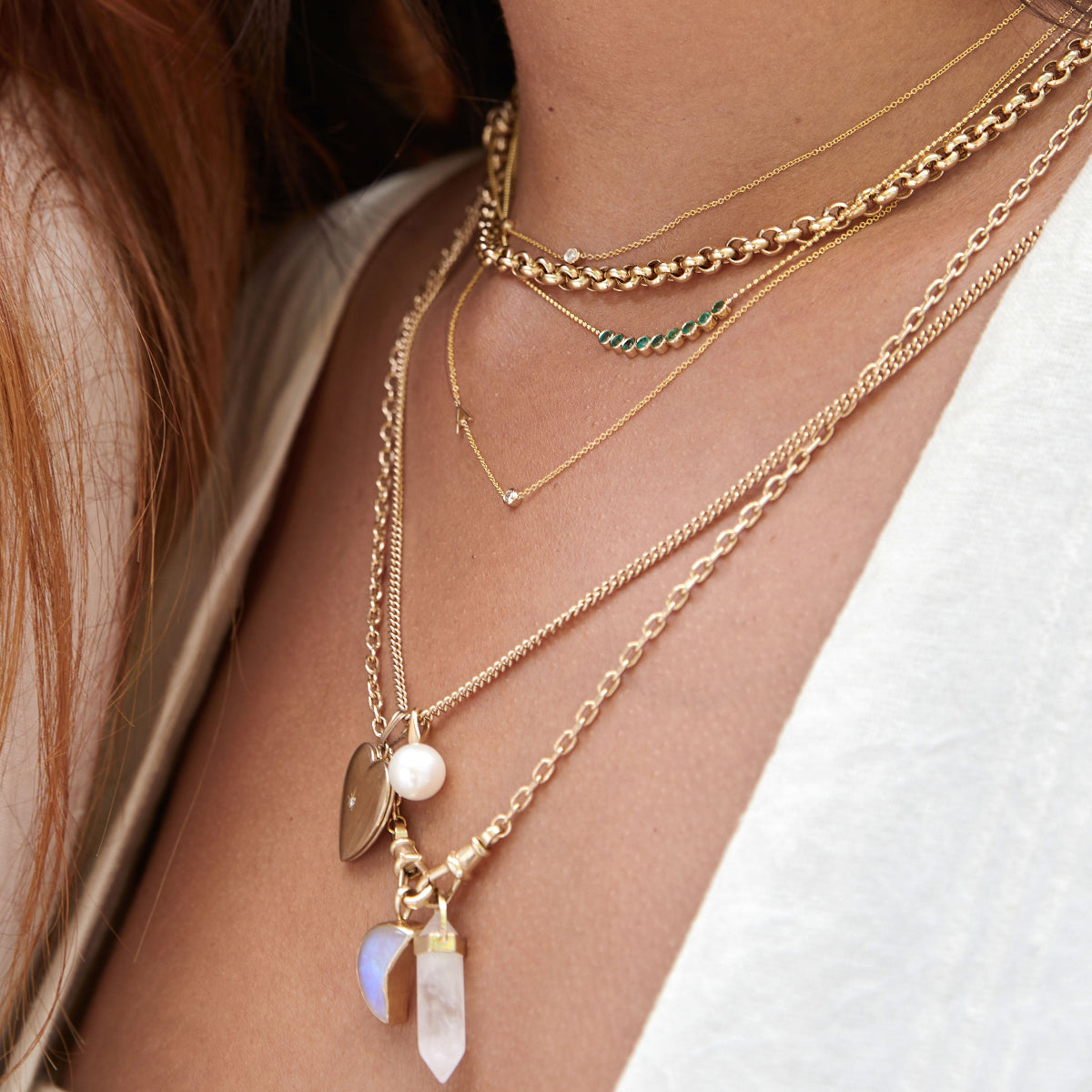 Close-up of a décolletage wearing a Rolo Necklace, a Diamond Layering Necklace, a Birthstone Arc Layering Necklace, a Monogram Necklace with Diamond, a Round Curb Chain with a Diamond Heart Locket Charm and a Perla Pendant, an Oval Link Chain with a Moonstone Crescent Charm and a Balance Crystal Charm, all in yellow gold, with a white top and some orange hair visible. 