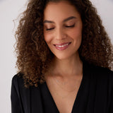 A woman wearing three layered Monogram Necklaces with Diamond charms, one in yellow gold with the letter M, one in white gold with the letter R and one in rose gold with the letter J, with brown curly hair and a black top, against a neutral background.