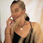 A woman wearing Endless Summer Necklaces in Sweet Escape and Santorini, The Ultimate Diamond Necklace with The Golden Venus Shell, Oval Hoops, an Endless Summer Bracelet - Coral Reef, an Element Short Link Bracelet and a yellow gold Simple Bangle, styled in a black strap top against a light background.