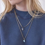 Close-up of a blonde woman's décolletage wearing The Thistle Bouquet Necklace, an Overboard Layering Chain with an Abalone Leaf Charm and a Box Chain with a Moonstone Crescent Charm, with a dark blue jumper against a light plain background. 