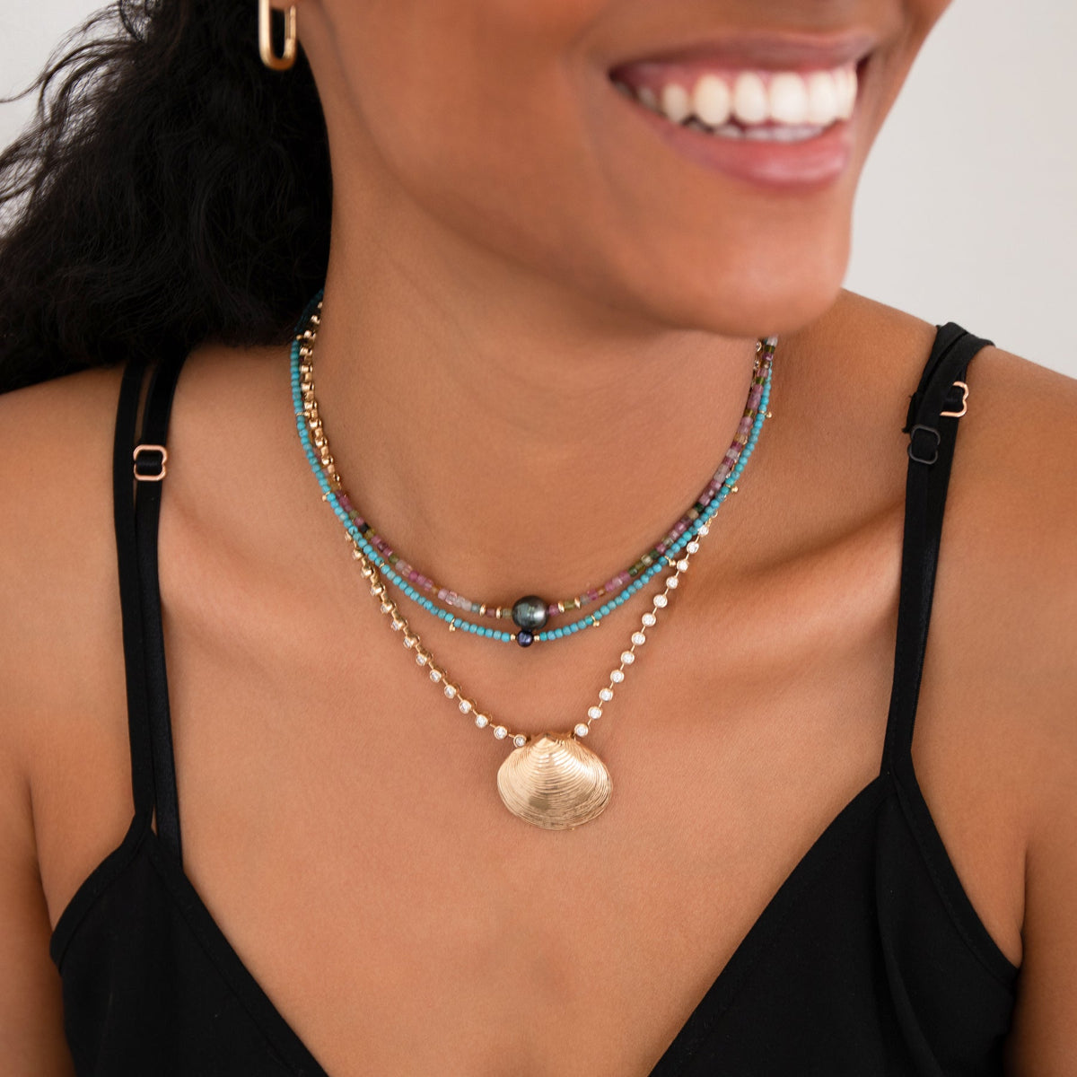 A close-up of a woman's neck wearing Endless Summer Necklaces in Sweet Escape and Santorini, The Ultimate Diamond Necklace with The Golden Venus Shell and Oval Hoops, styled in a black strap top. 