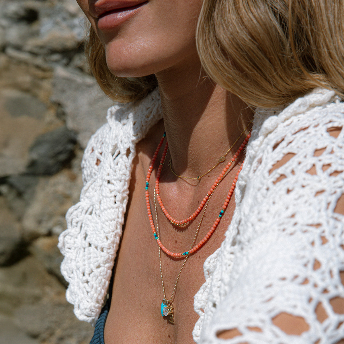 Close-up of a blonde woman's neck wearing a yellow gold Custom Gold Necklace – 4 Letters, Endless Summer Necklaces in Mykonos and Calypso, a yellow-gold Classic Bead Chain with an Etched Turquoise Heart Charm, a Diamond Pendant and a Petite Nameplate Pendant, styled in a white crochet top against a stone-blurred background.